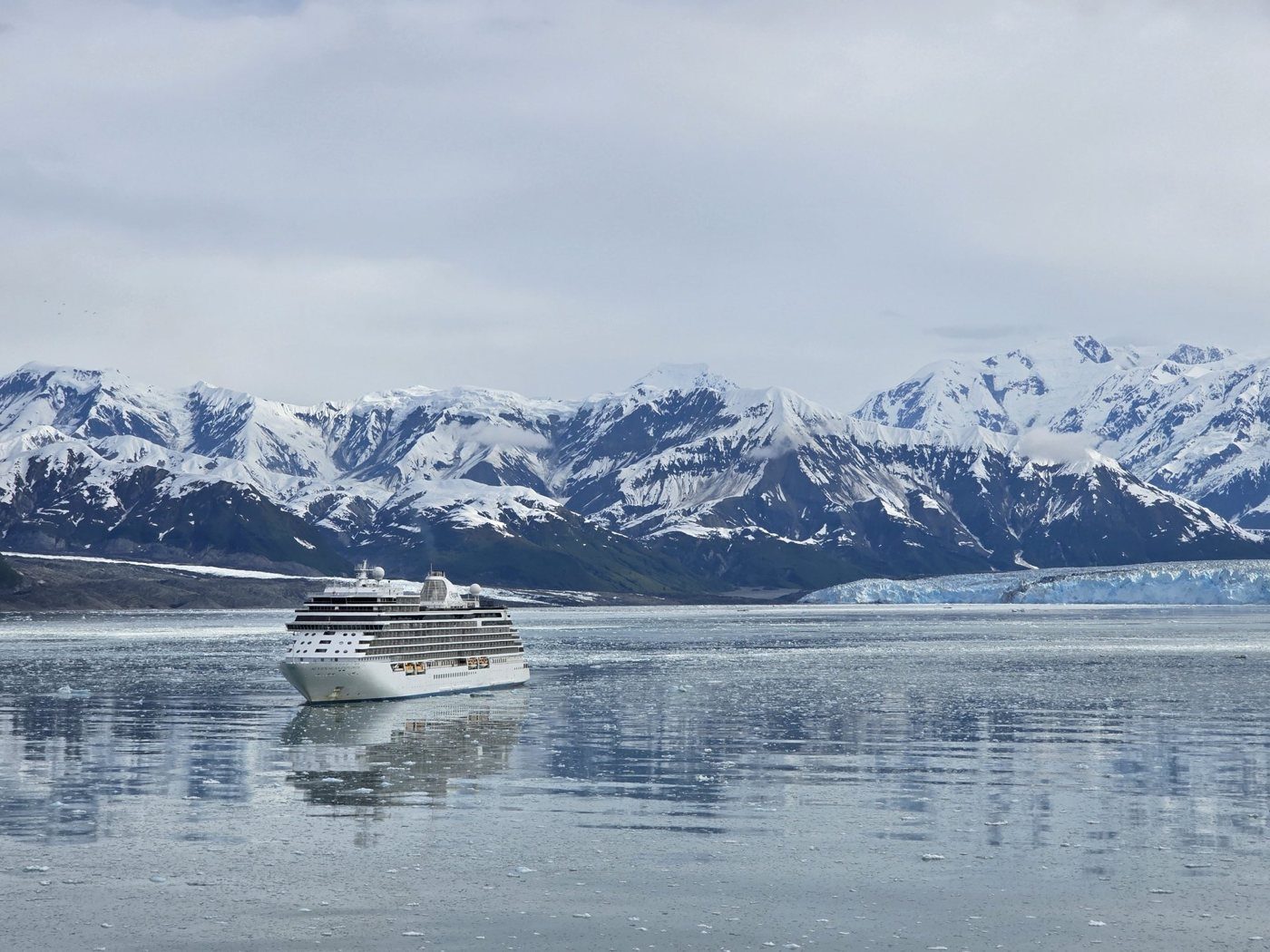 A small ship sailing toward a glacier beneath snowy mountains